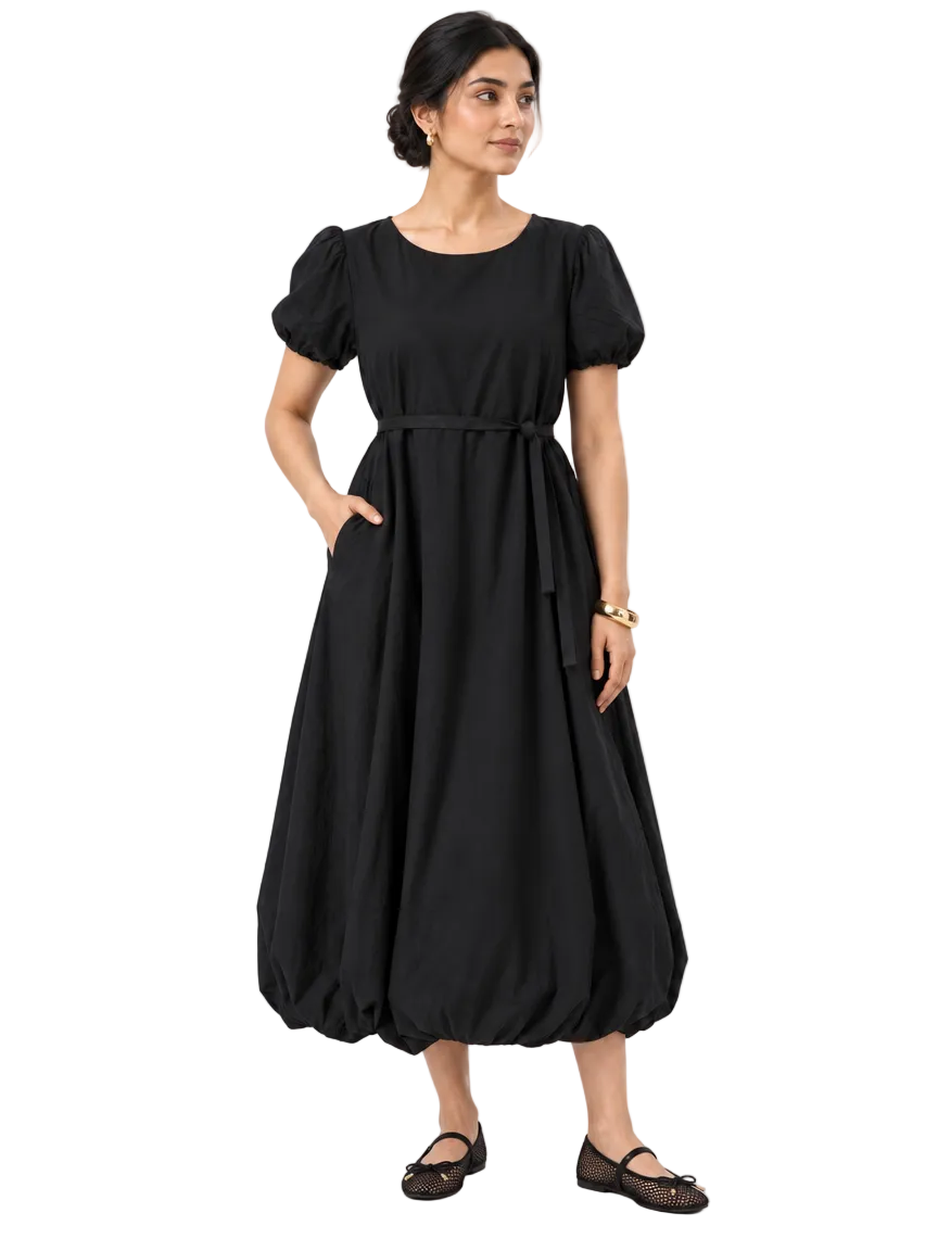 Front view of The Lamorah black puff-sleeve balloon-hem dress with a narrow matching sash, styled with black mesh Mary Jane flats and a gold cuff bracelet.