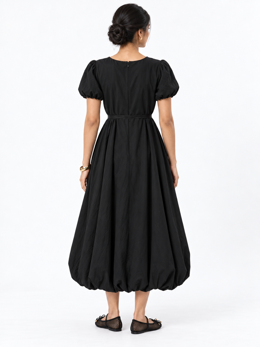 Back view of The Lamorah black balloon-hem dress showing its centre-back zipper, puff sleeves and narrow matching sash, styled with mesh flats.