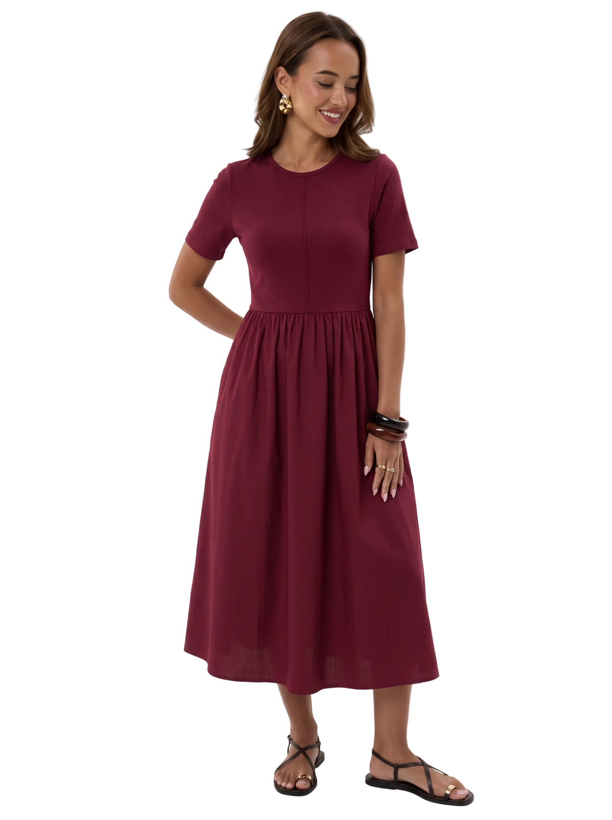 Smiling model wearing The Lamorah burgundy stretch-bodice gathered midi dress, with hands out of pockets to show the full skirt, styled with flat sandals and chunky bangles.