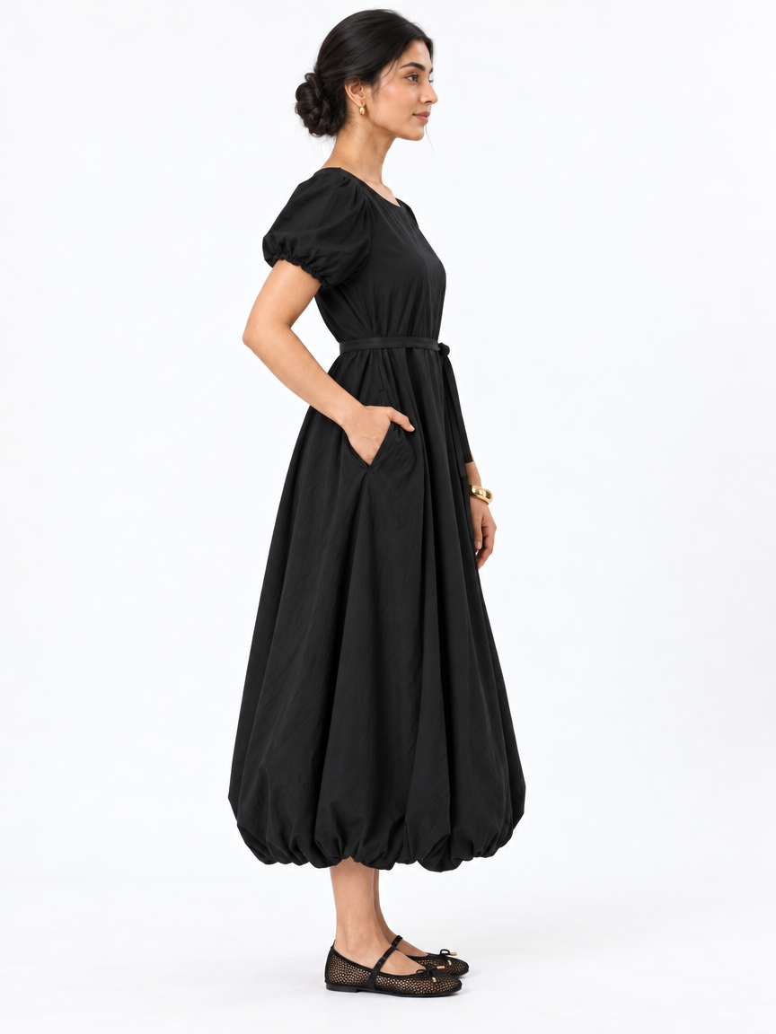 Side profile of The Lamorah black puff-sleeve dress showing its pocket, narrow sash and rounded balloon hem, styled with black mesh Mary Jane flats.