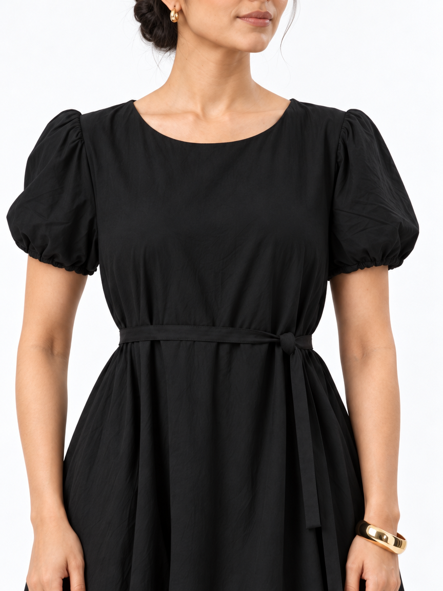 Close-up of The Lamorah black dress showing its round neckline, puff sleeves and sleeve openings, narrow sash knot, fabric texture and styled gold cuff bracelet.