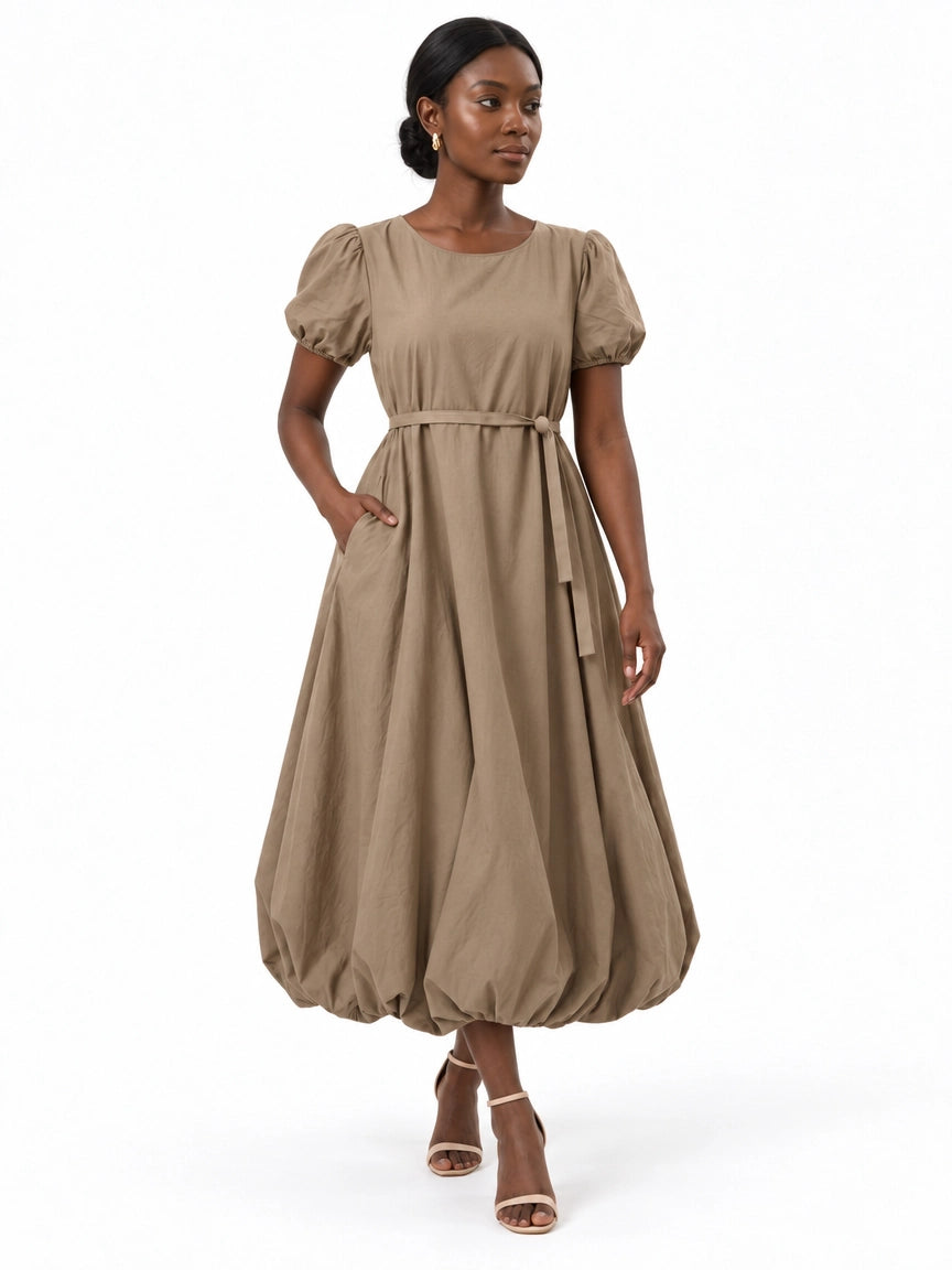 Walking front view of The Lamorah taupe balloon-hem dress with puff sleeves, side pocket and a narrow matching waist sash.
