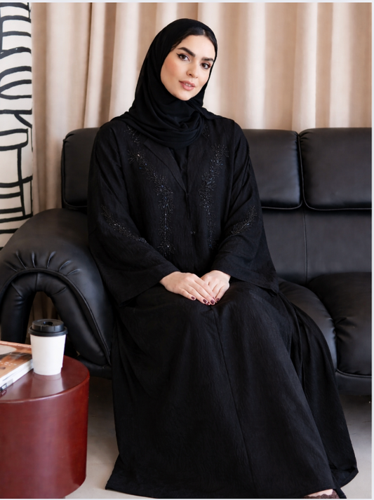 Woman in a black abaya sitting on a black couch with a beige curtain in the background |THE LAMORAH
