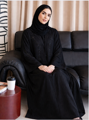Woman in a black abaya sitting on a black couch with a beige curtain in the background |THE LAMORAH
