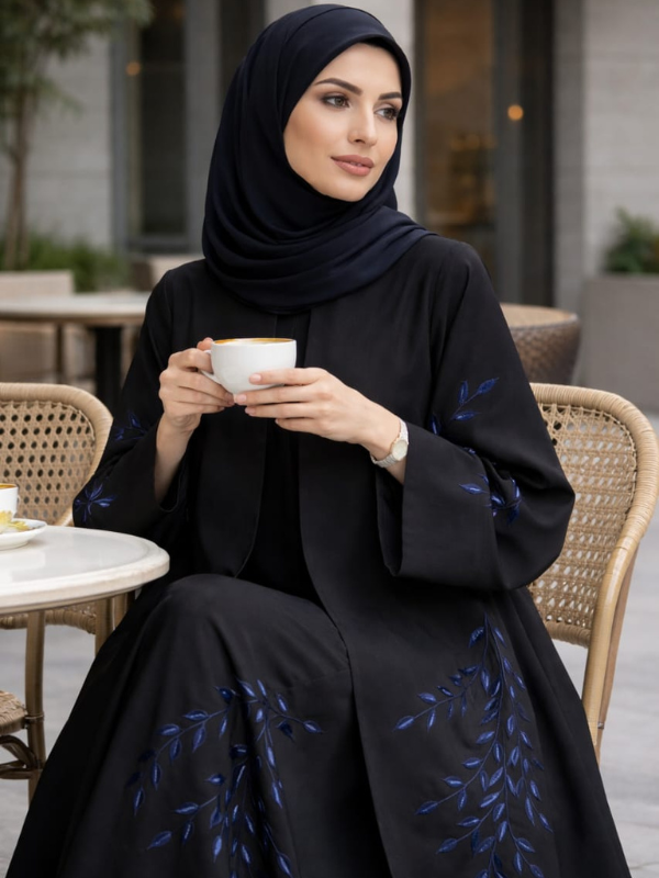 Woman in a black abaya with blue embroidery holding a cup outdoors.|THE LAMORAH
