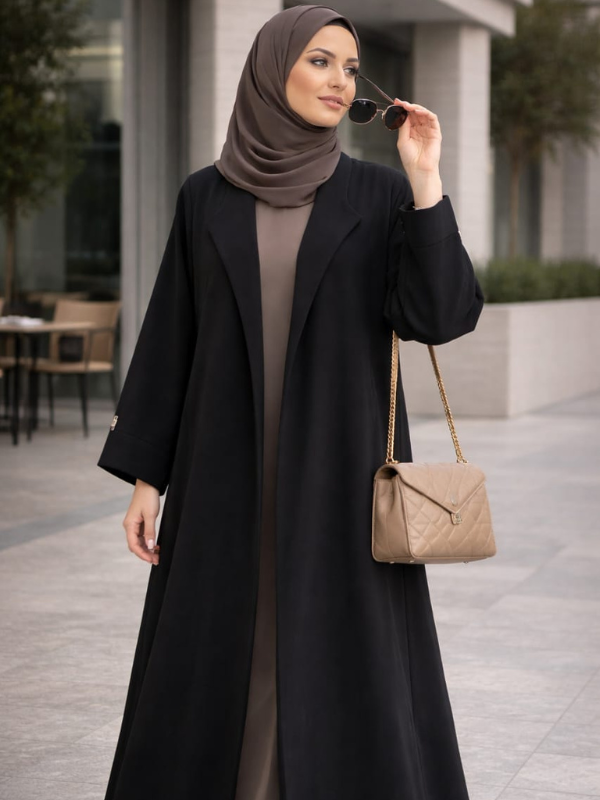 Woman in a black coat and hijab holding sunglasses, standing outdoors. |THE LAMORAH
