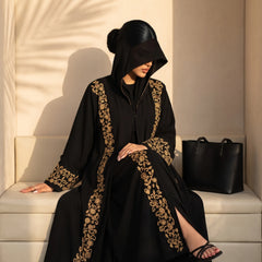 Black hooded abaya with antique gold scrolling embroidery down the front panels and cuffs, worn seated